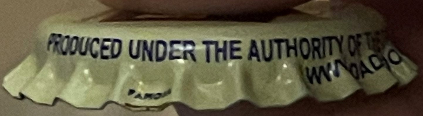 Vintage bottle cap with text "PRODUCED UNDER THE AUTHORITY OF" and partial text visible, shown at angle against tan backgroun