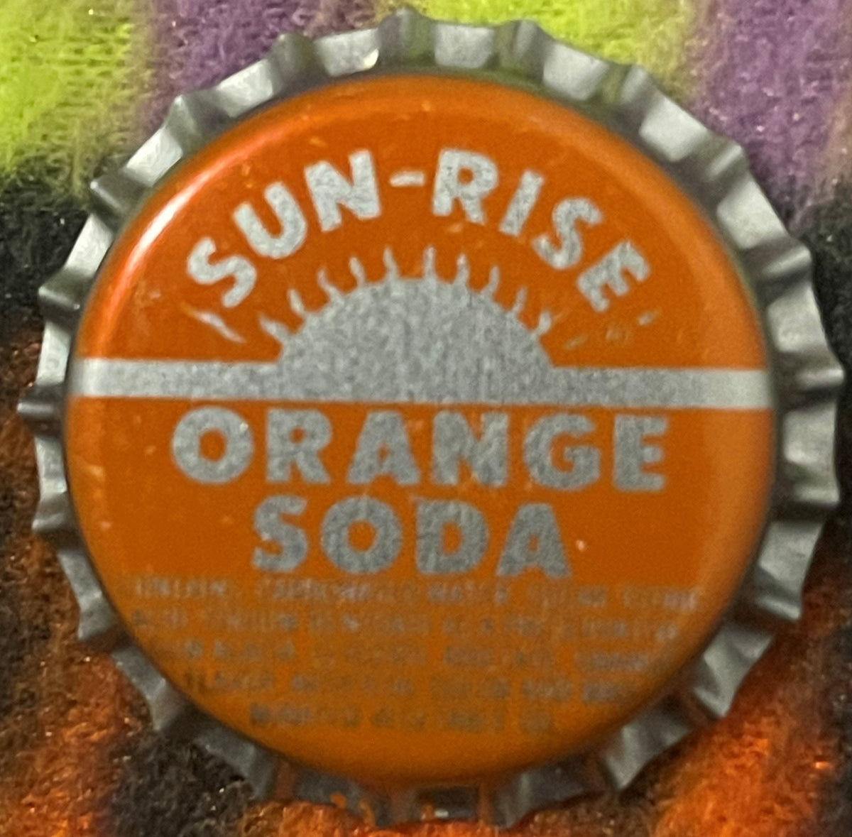 Vintage orange bottle cap with white ridged edge, printed text reading "SUN-RISE ORANGE SODA" and a sun design, photographed 