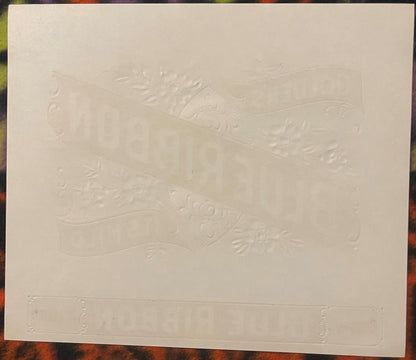 Embossed cream-colored cigar label with decorative scrollwork and ornamental border, shown flat on patterned fabric backgroun