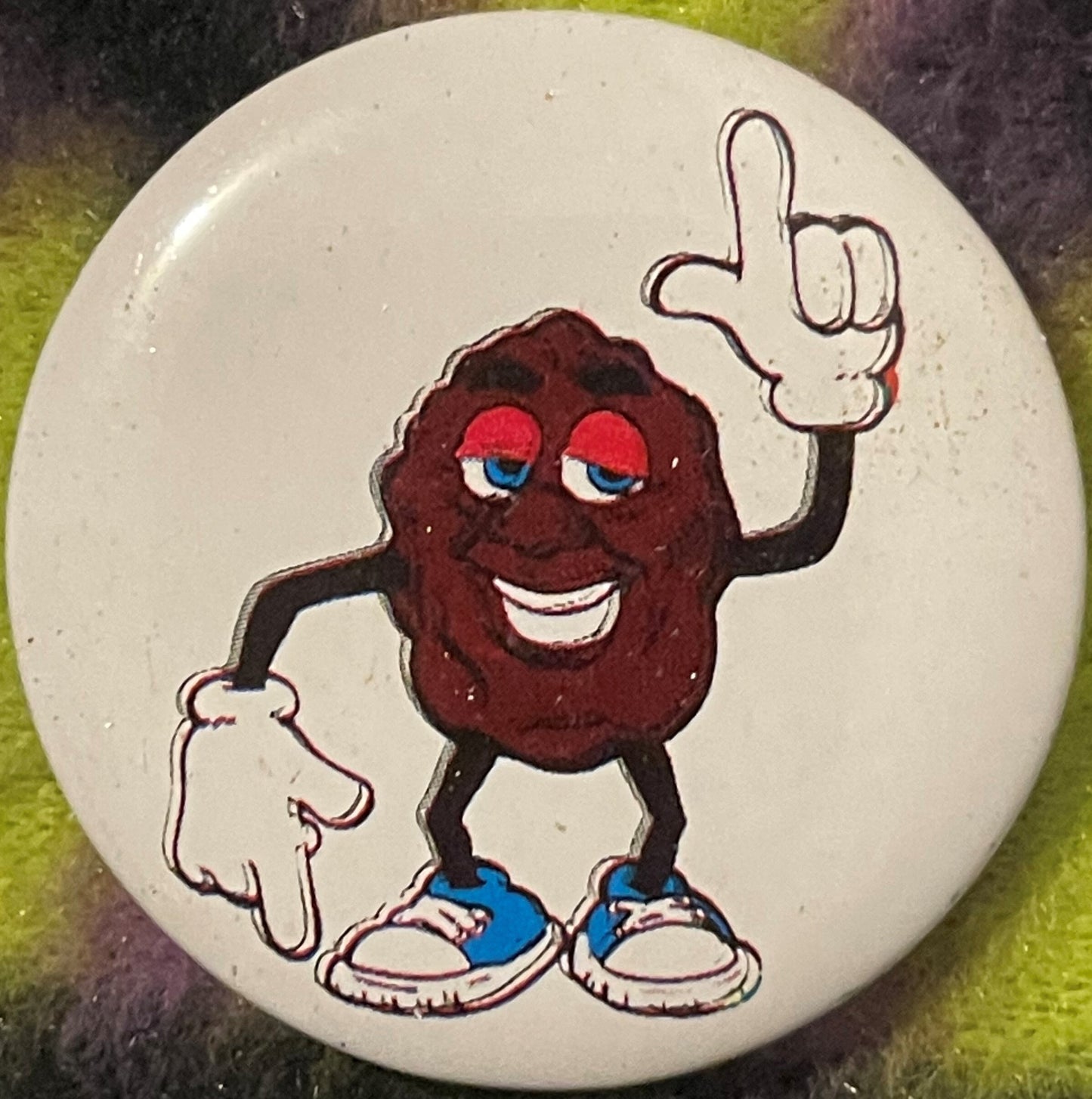 Close-up of a cream-colored pinback button featuring a smiling dark brown raisin character with red eyes, white gloves, and b