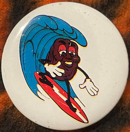 Round white pinback button featuring a cartoon character surfing on a blue wave, wearing red and yellow striped shorts, with 
