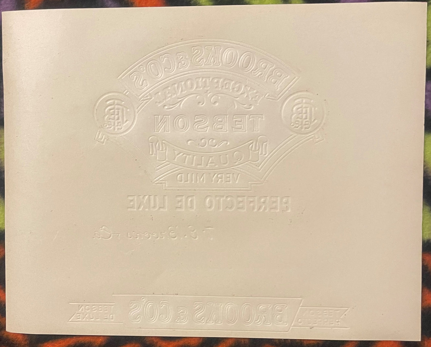 Cream-colored embossed cigar label with ornate oval badge design and reversed text, photographed flat against colored backgro