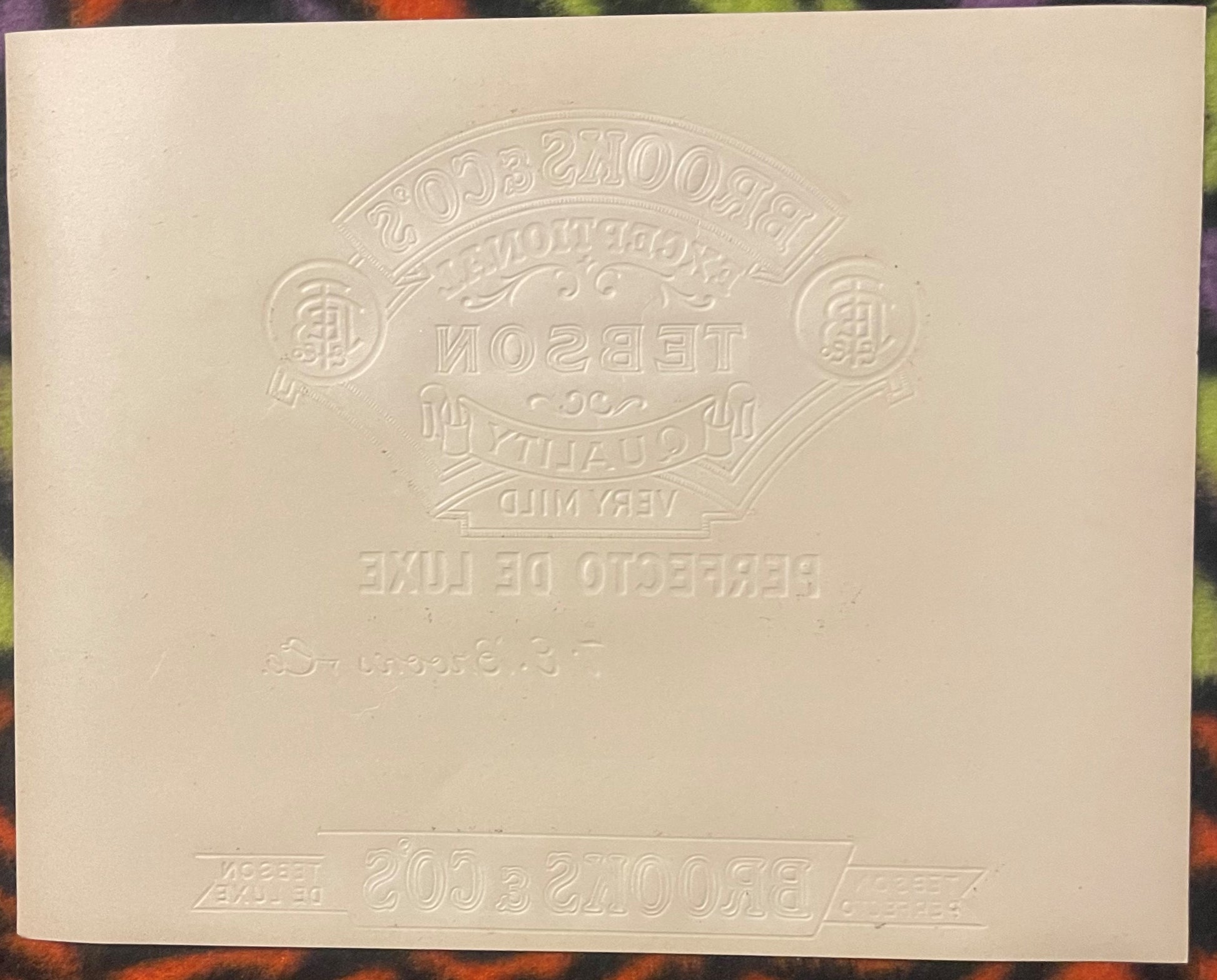 Cream-colored embossed cigar label with ornate oval badge design and reversed text, photographed flat against colored backgro