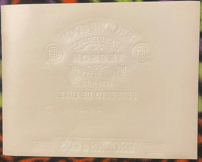 Cream-colored embossed cigar label with ornate oval badge design and reversed text, photographed flat against colored backgro