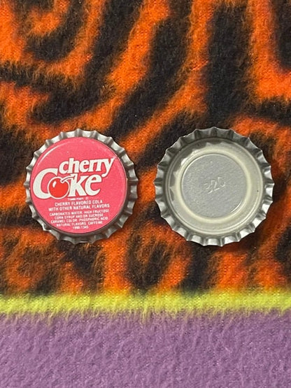 Vintage Cherry Coke bottle cap showing red front with logo and silver metallic back, displayed on colorful patterned fabric