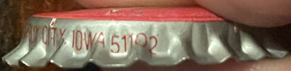 Close-up of vintage bottle cap with red top and white ridged sides, held by a finger for scale, showing partial red text and 