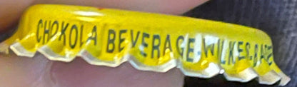 Vintage yellow bottle cap with scalloped edges held in hand, printed text reads "CHOKOLA BEVERAGE" and "WILKES-BARRE PA