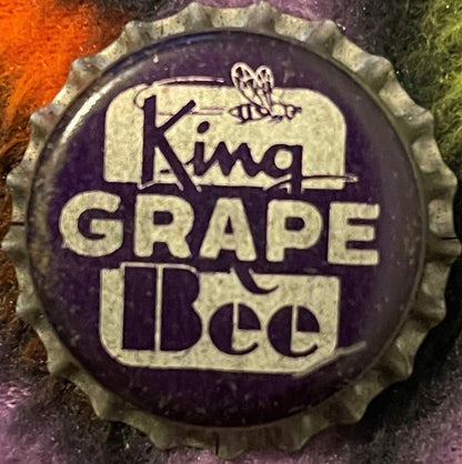 Antique King Bee Grape Soda bottle cap, cork-lined crown with embossed bee illustration and purple and cream text on dark background.
