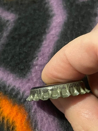 Close-up profile of the King Bee cap's crimped edge and embossed rim text ending "...DRATE OF SODA ARTIFICIAL FLAVOR...", held by fingertip against striped fabric