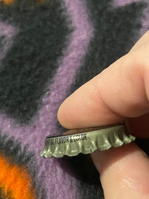 Close-up profile of the King Bee cap's crimped edge and embossed rim text ending "...FICIAL FLAVOR & COLOR", held between fingers against fabric