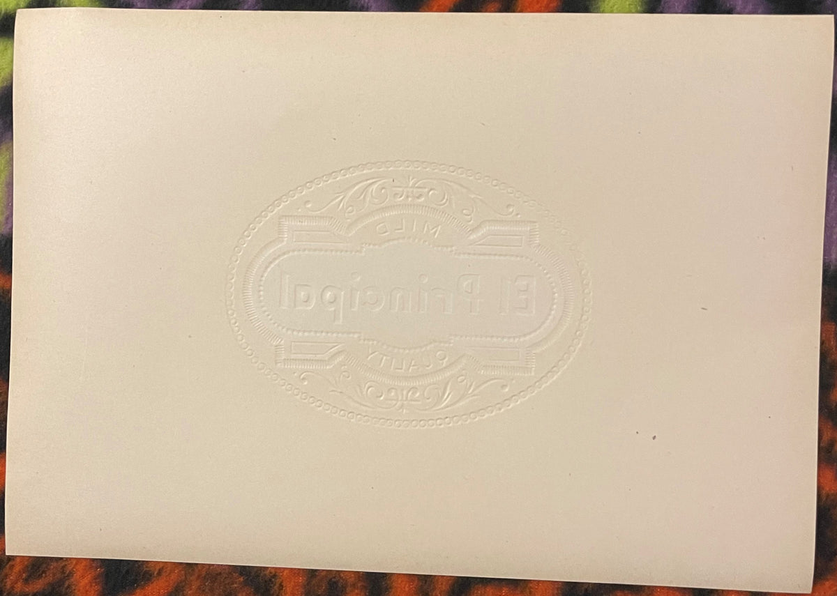 Cream-colored embossed cigar label with ornate oval border and "El Principal" text, displayed flat on colorful fabric