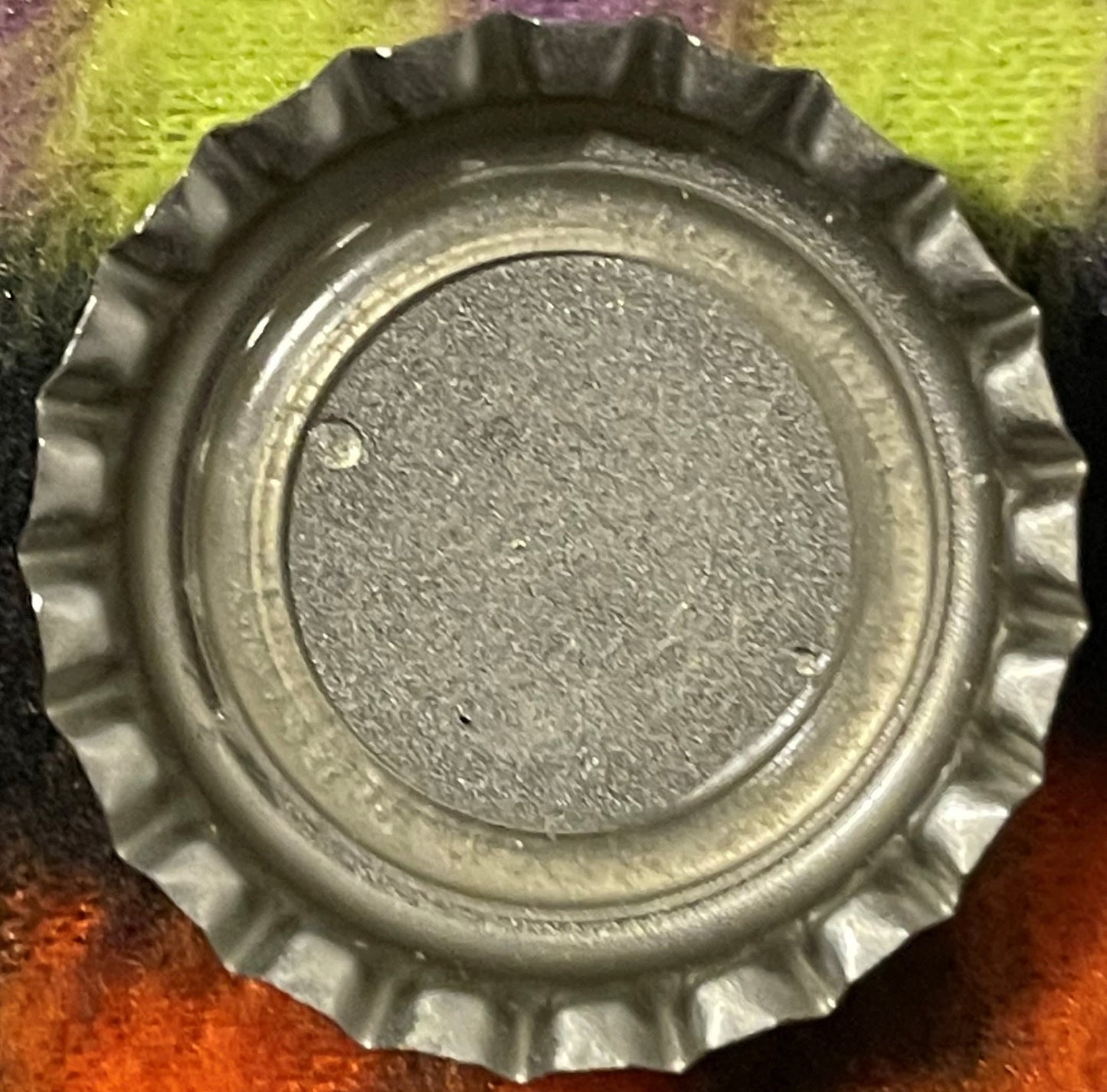 Vintage metal bottle cap with ridged crown edges, shown flat from above on colored background, surface shows wear and patina