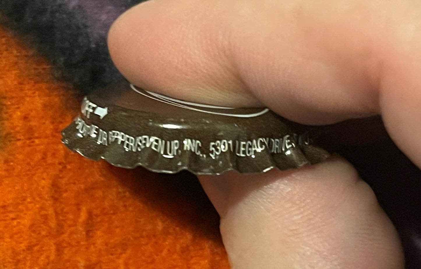 Close-up of a metal twist-off bottle cap held by a thumb, showing embossed text referencing A&W Root Beer and West Jefferson,