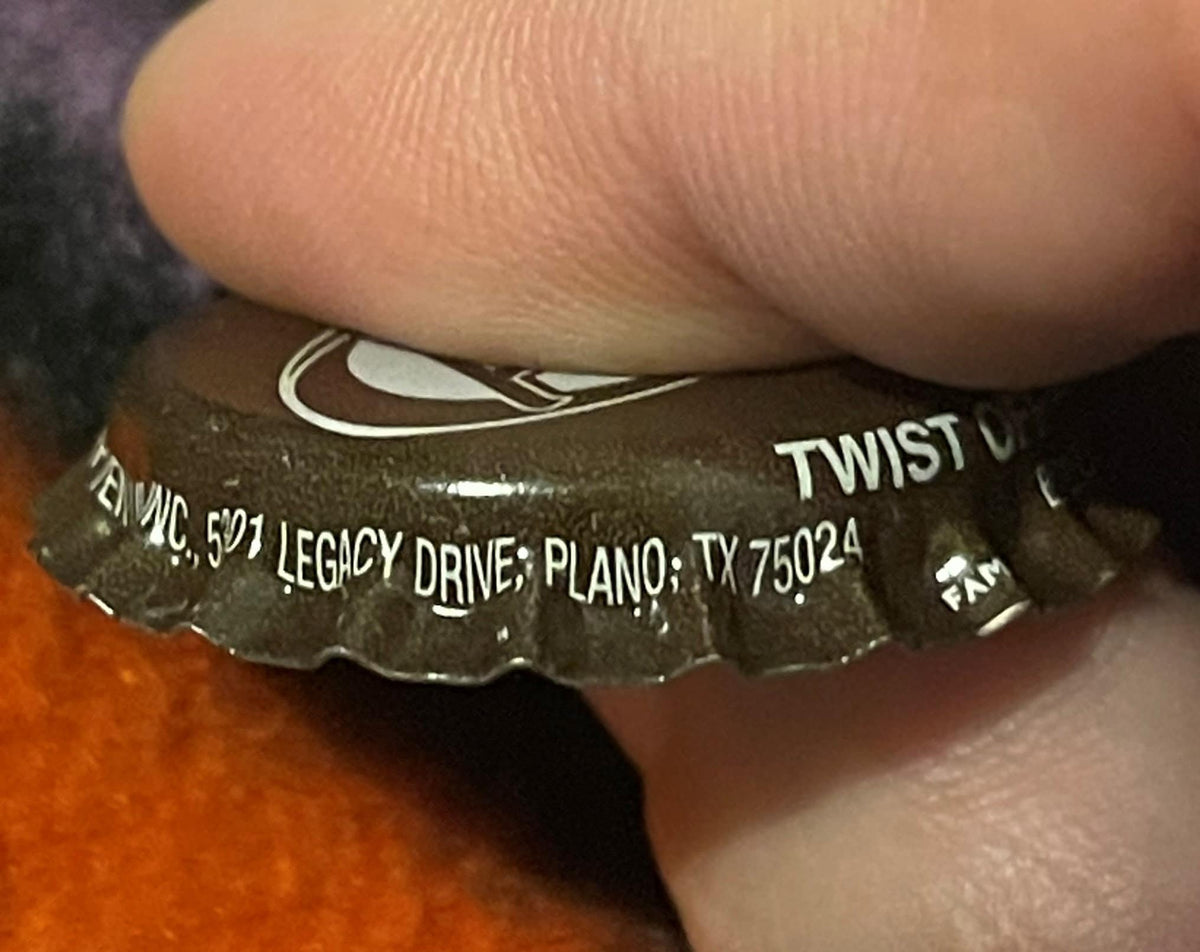 Close-up of brown twist-off bottle cap held by thumb, showing Nike swoosh logo and text reading "LEGACY DRIVE; PLANO; TX 7502