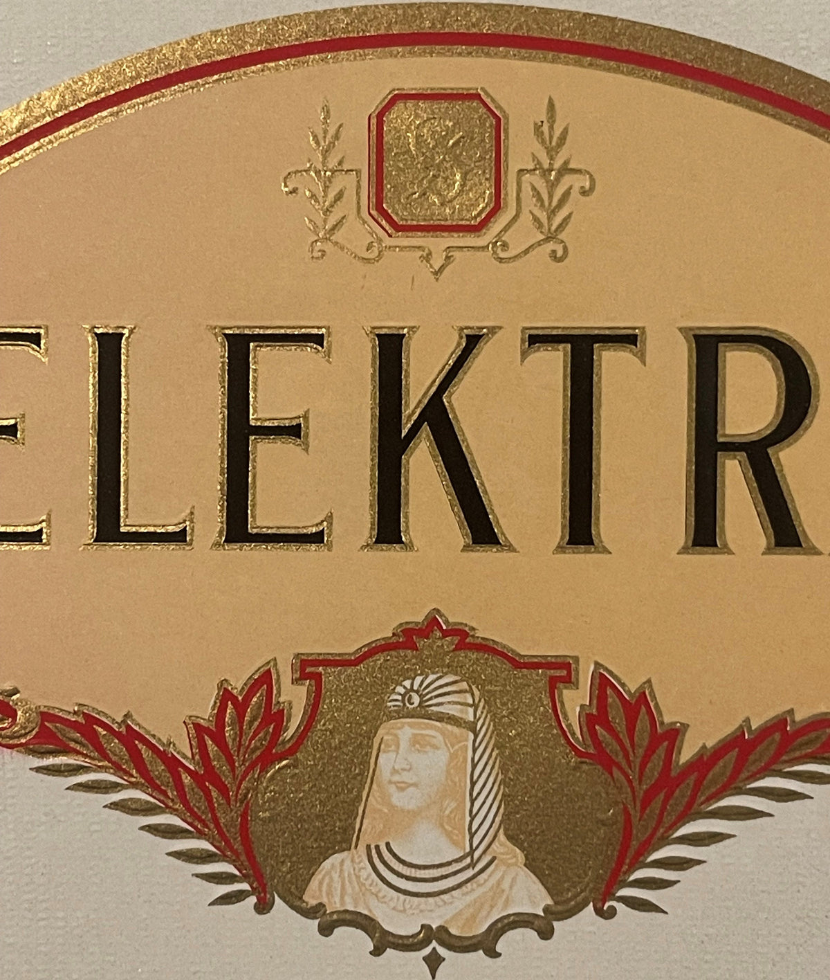 Close-up of the Elektra cigar label: ELEKTRA in raised gold-outlined letters, the script monogram in its red-and-gold octagon, and the woman in a striped Egyptian-style headdress framed in red and gold leaves