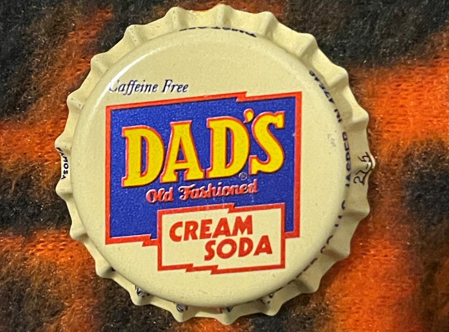 Vintage cream-colored bottle cap with blue and red "Dad's Old Fashioned Cream Soda" label, shown flat against a mottled rust 