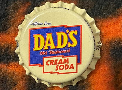 Vintage cream-colored bottle cap with blue and red "Dad's Old Fashioned Cream Soda" label, shown flat against a mottled rust 