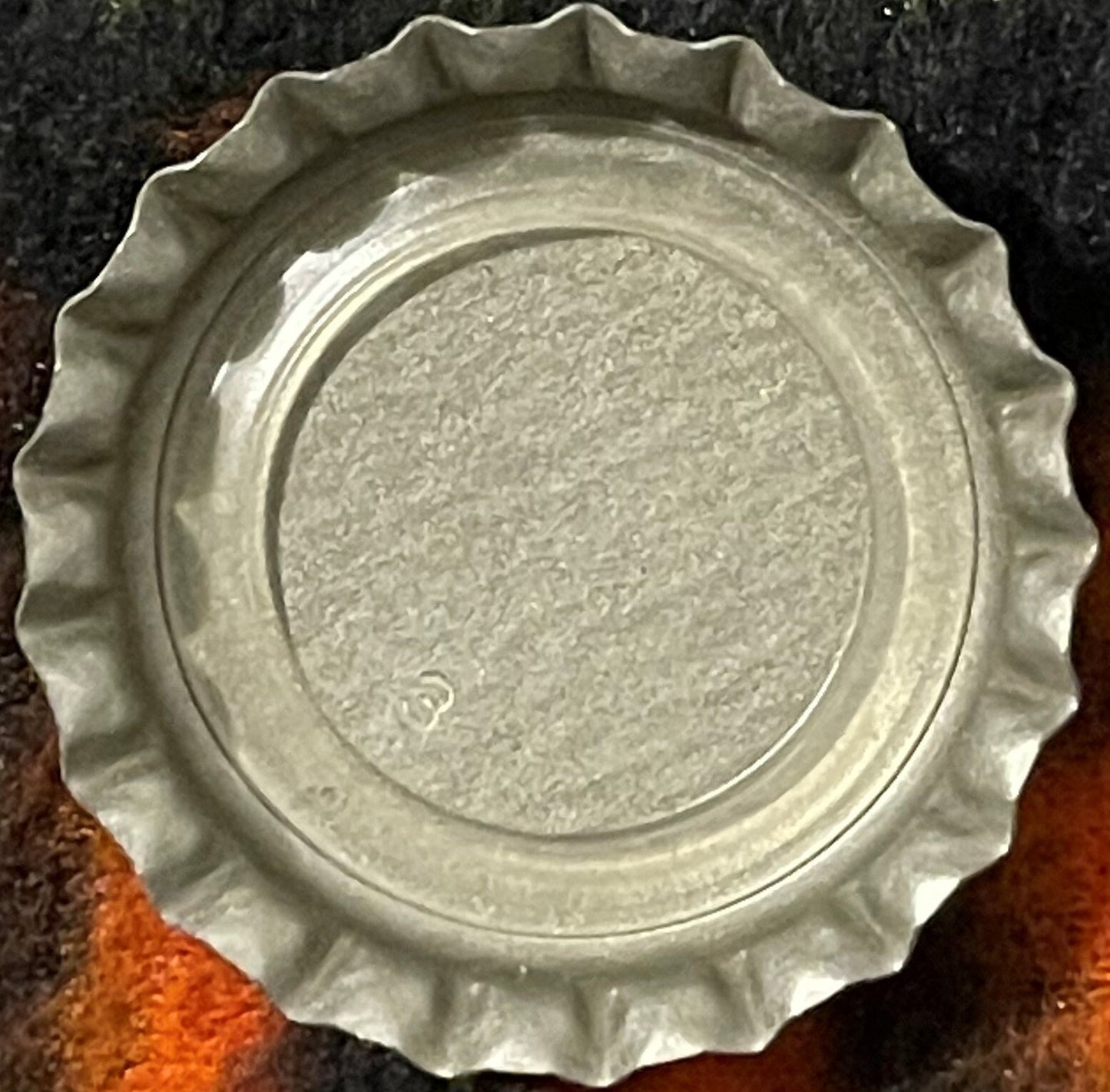 Vintage cream soda bottle cap with ridged metal edges and embossed center, photographed flat overhead on orange and dark fabr