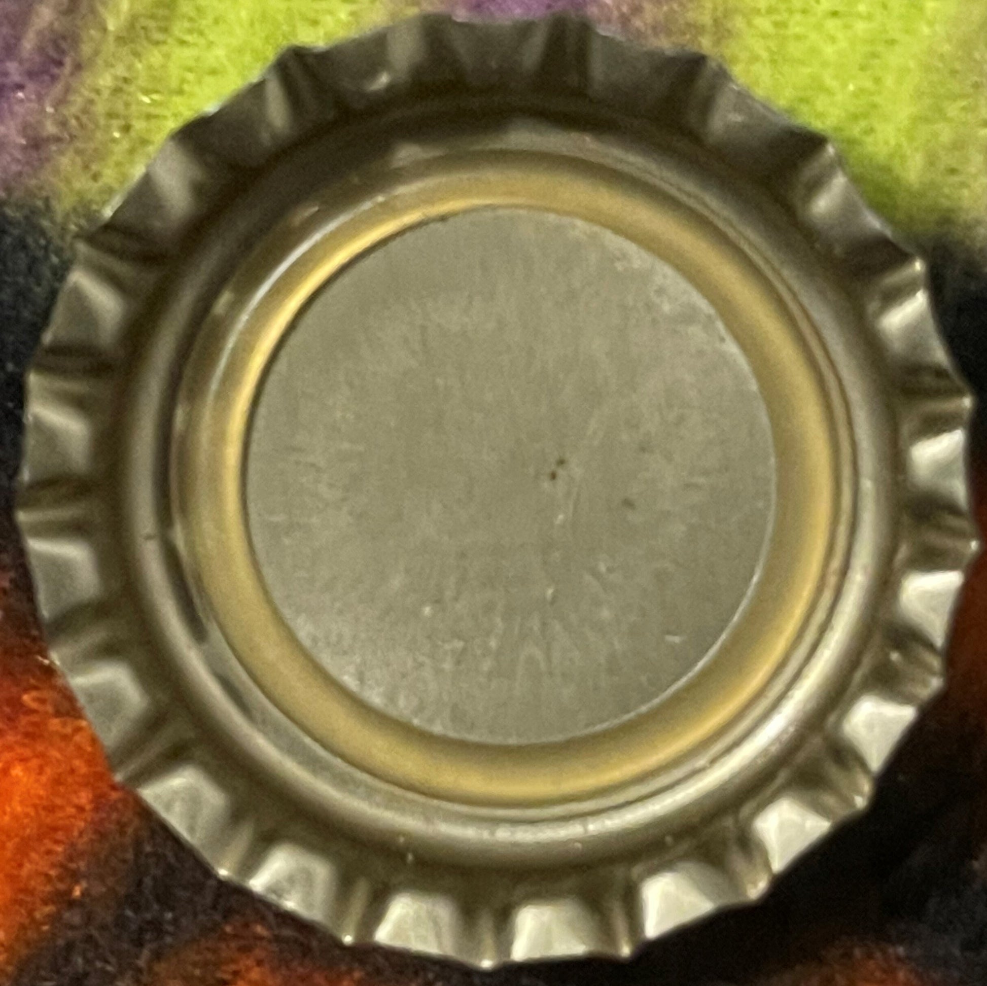 Vintage metal bottle cap with crimped edges and plain center, photographed from above on colorful fabric background