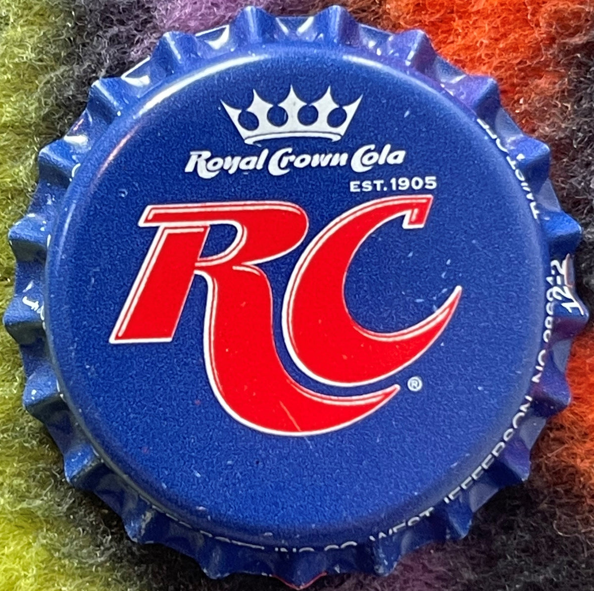 Blue Royal Crown Cola bottle cap with red RC logo and crown, "EST. 1905" text visible, on colorful textile background