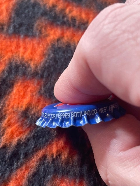 Vintage blue Royal Crown Cola bottle cap held in fingertips, showing embossed text referencing Dr Pepper Bottling Co West