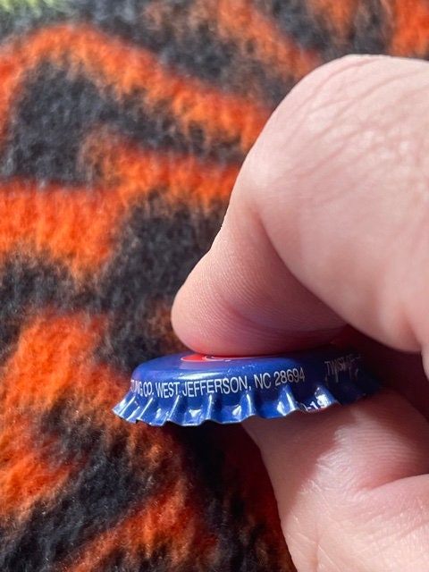 Blue Royal Crown Cola bottle cap held in fingers, showing "West Jefferson NC" text, with orange and black patterned fabric