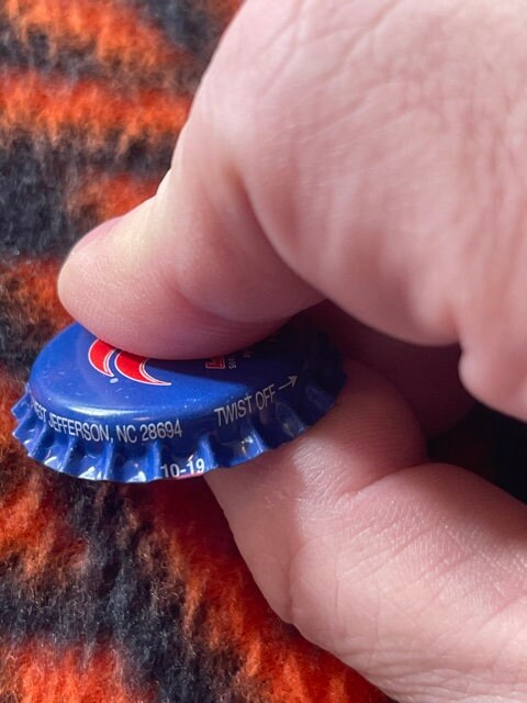 Close-up of blue RC Cola bottle cap held in fingers, showing red crown logo and West Jefferson NC text for scale.