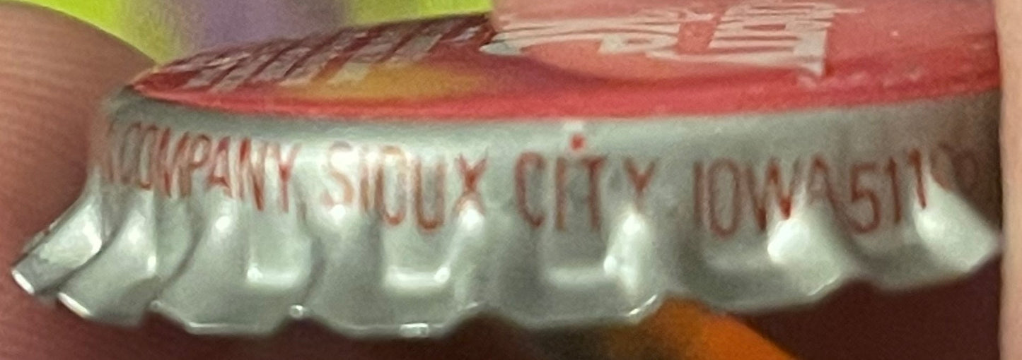 Close-up of a ridged bottle cap with red and white printed text reading "COMPANY SIOUX CITY IOWA" against a blurred reddish b