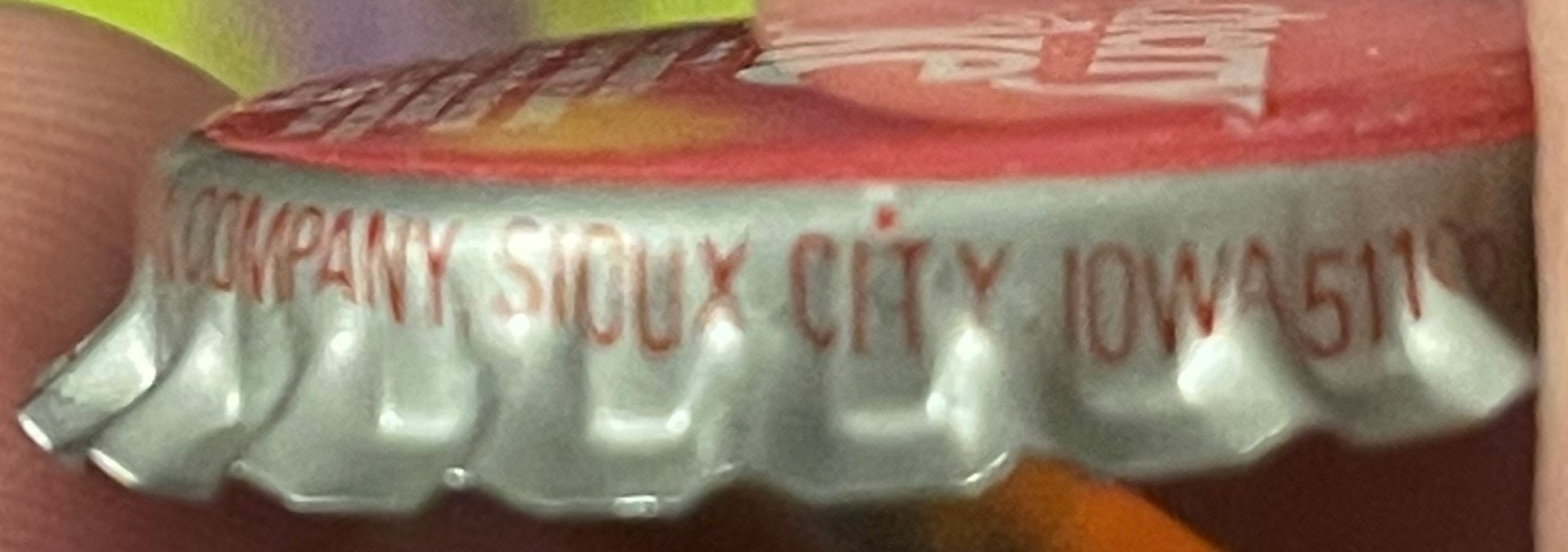 Close-up of a ridged bottle cap with red and white printed text reading "COMPANY SIOUX CITY IOWA" against a blurred reddish b