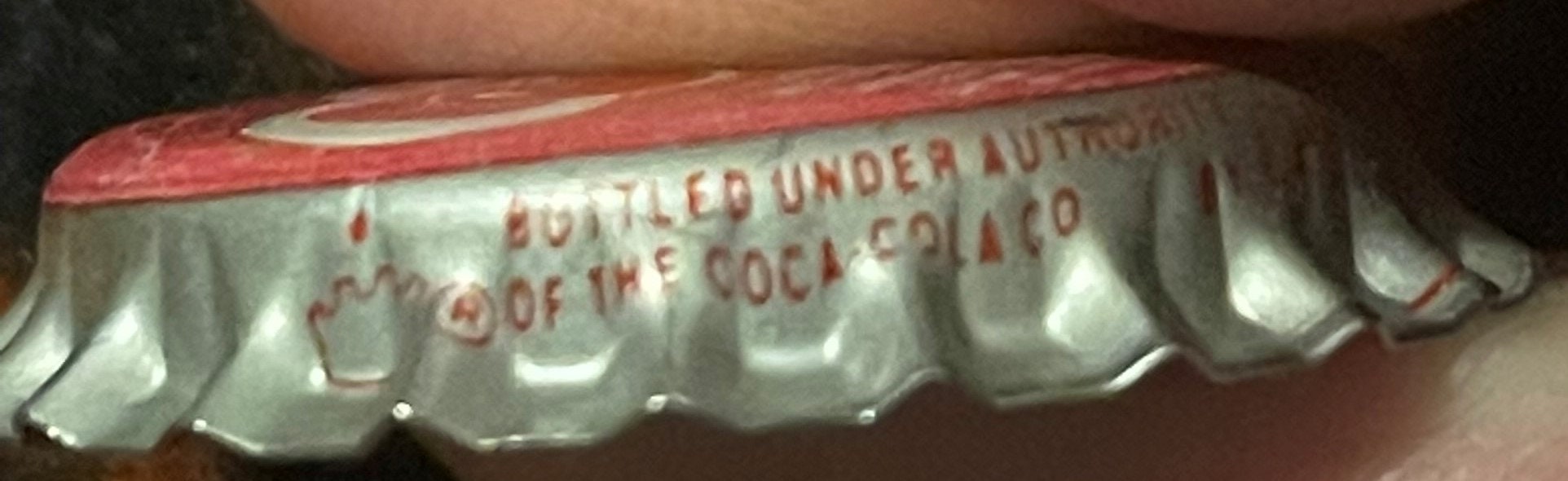 Close-up of red and white bottle cap held by a finger, showing embossed text "BOTTLED UNDER AUTHORITY OF THE COCA-COLA CO