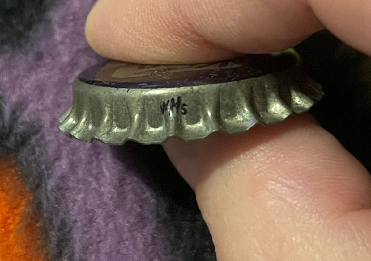 Close-up profile of the King Bee cap's crimped metal edge, held between thumb and finger, with a handwritten ink mark reading "WMS" on the rim