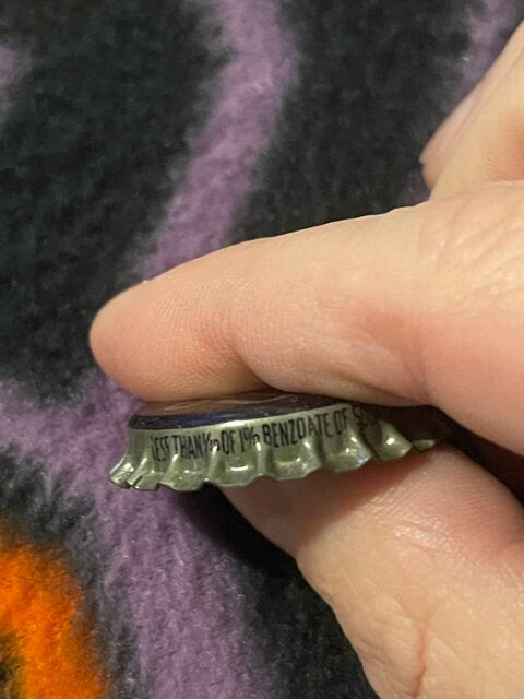 Close-up of antique King Bee Grape Soda crown bottle cap held between thumb and finger, showing embossed text and ridged metal edge against colorful fabric background.