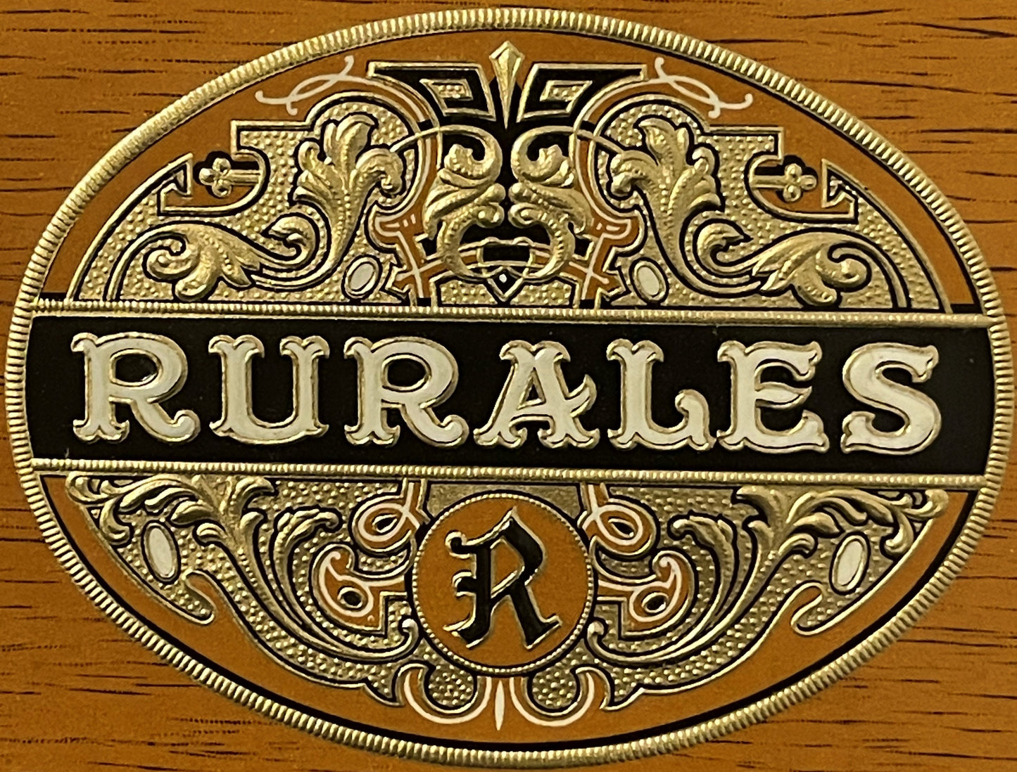 Embossed brass cigar label with ornate scrollwork reading "RURALES" on black band, mounted on wood grain background