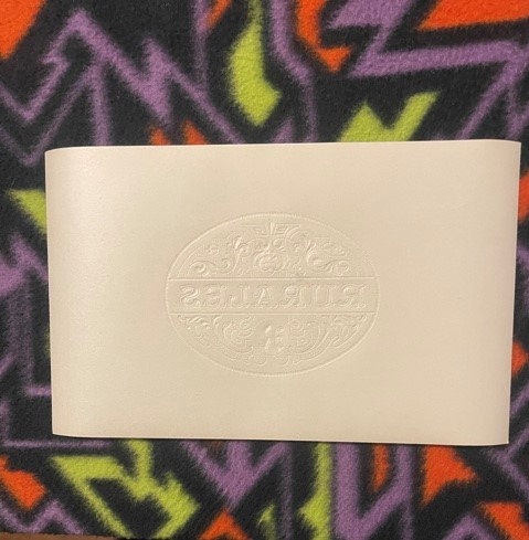 Cream-colored embossed cigar label with oval "Rurales" text, displayed against colorful patterned fabric background