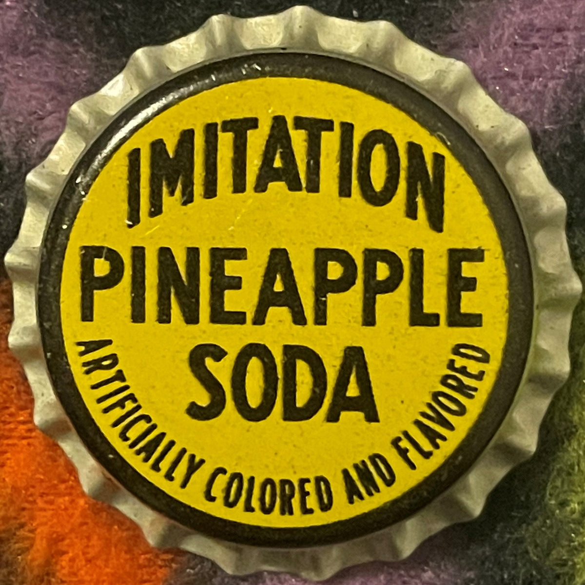 Imitation Pineapple Soda cork bottle cap, front: yellow face in a black ring with black block letters and ARTIFICIALLY COLORED AND FLAVORED along the lower edge, fluted silver-toned skirt