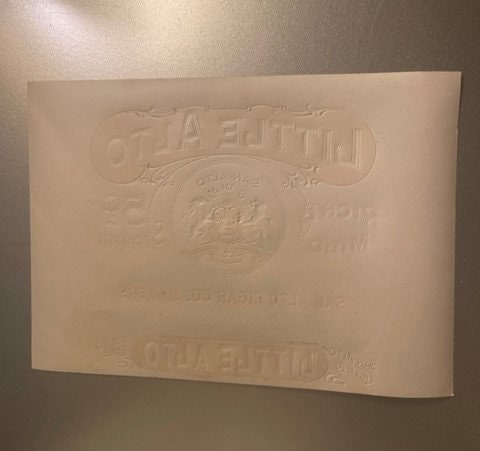 Cream-colored embossed cigar label with "Little Alto" text at top and bottom, photographed at angle against brown wall