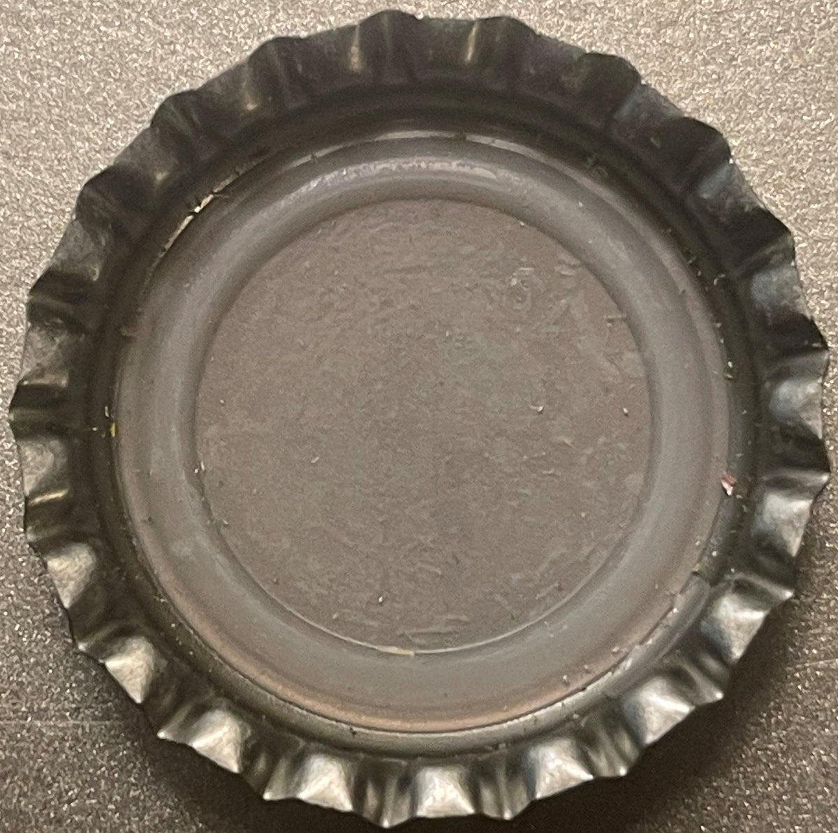 Overhead view of a metallic bottle cap with crimped ridged edges and flat center, photographed on a textured gray surface