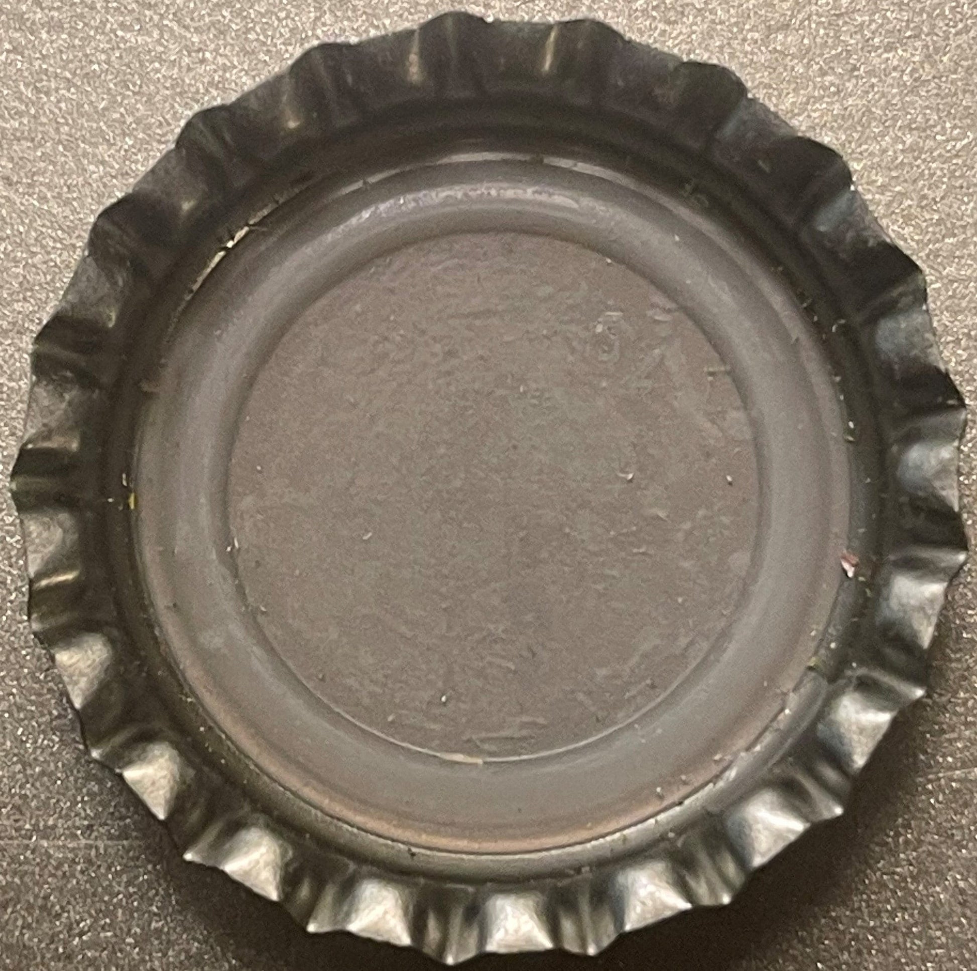 Overhead view of a metallic bottle cap with crimped ridged edges and flat center, photographed on a textured gray surface
