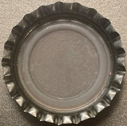 Overhead view of a metallic bottle cap with crimped ridged edges and flat center, photographed on a textured gray surface