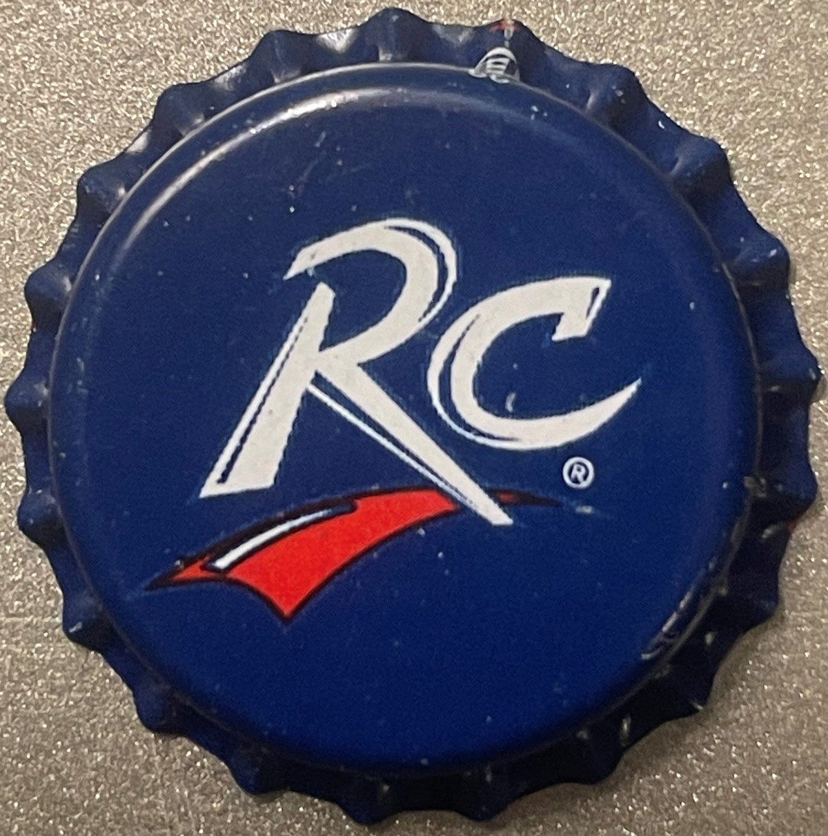 Navy blue bottle cap with white "RC" logo and red swoosh, shown flat against textured gray surface