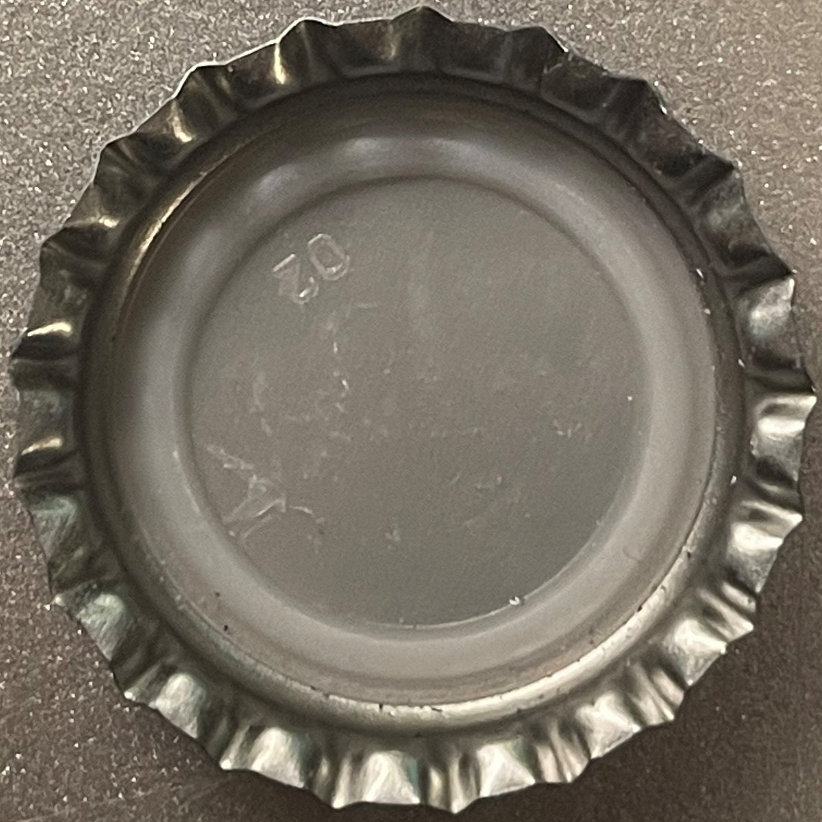 Metallic bottle cap with ridged crown edge viewed from above on gray surface, showing embossed text and numbers
