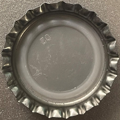 Metallic bottle cap with ridged crown edge viewed from above on gray surface, showing embossed text and numbers