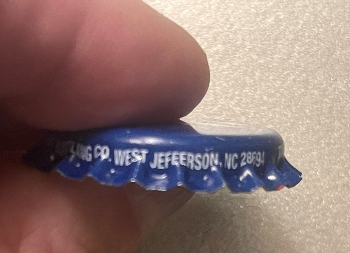 Blue metal bottle cap held by a finger, showing embossed text "West Jefferson, NC 28691