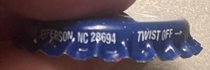 Blue metal bottle cap with "West Jefferson, NC 28694" and "TWIST OFF" text visible, held in hand for scale