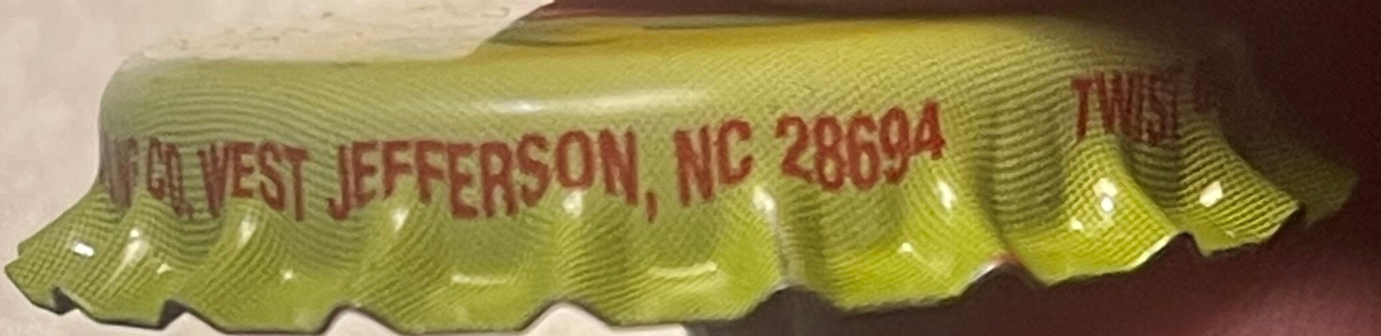 Close-up of yellow bottle cap with red embossed text reading "HARVEST JEFFERSON, NC 28694" and partial NASCAR-era branding vi