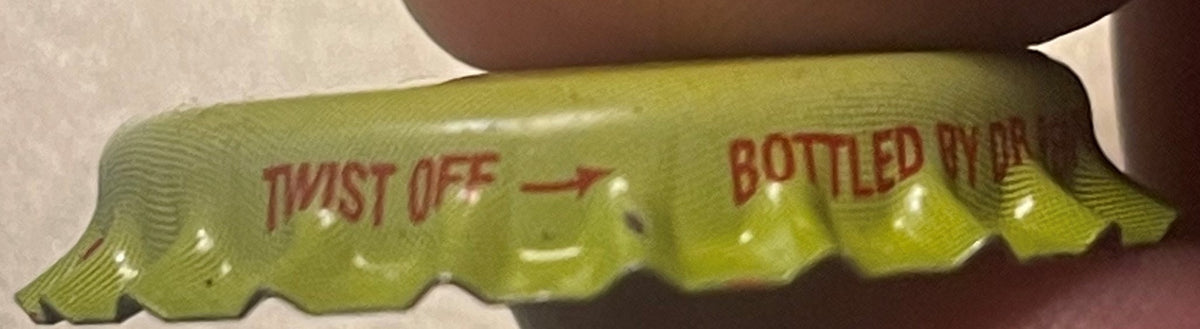 Vintage yellow-green Sun Drop bottle cap with red "TWIST OFF" and "BOTTLED BY" text, held in brown-gloved hand, close-up view