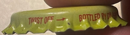 Vintage yellow-green Sun Drop bottle cap with red "TWIST OFF" and "BOTTLED BY" text, held in brown-gloved hand, close-up view