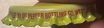 Yellow-green vintage bottle cap with red embossed text reading "Dr Pepper Bottling Co West" shown at angle against brown back