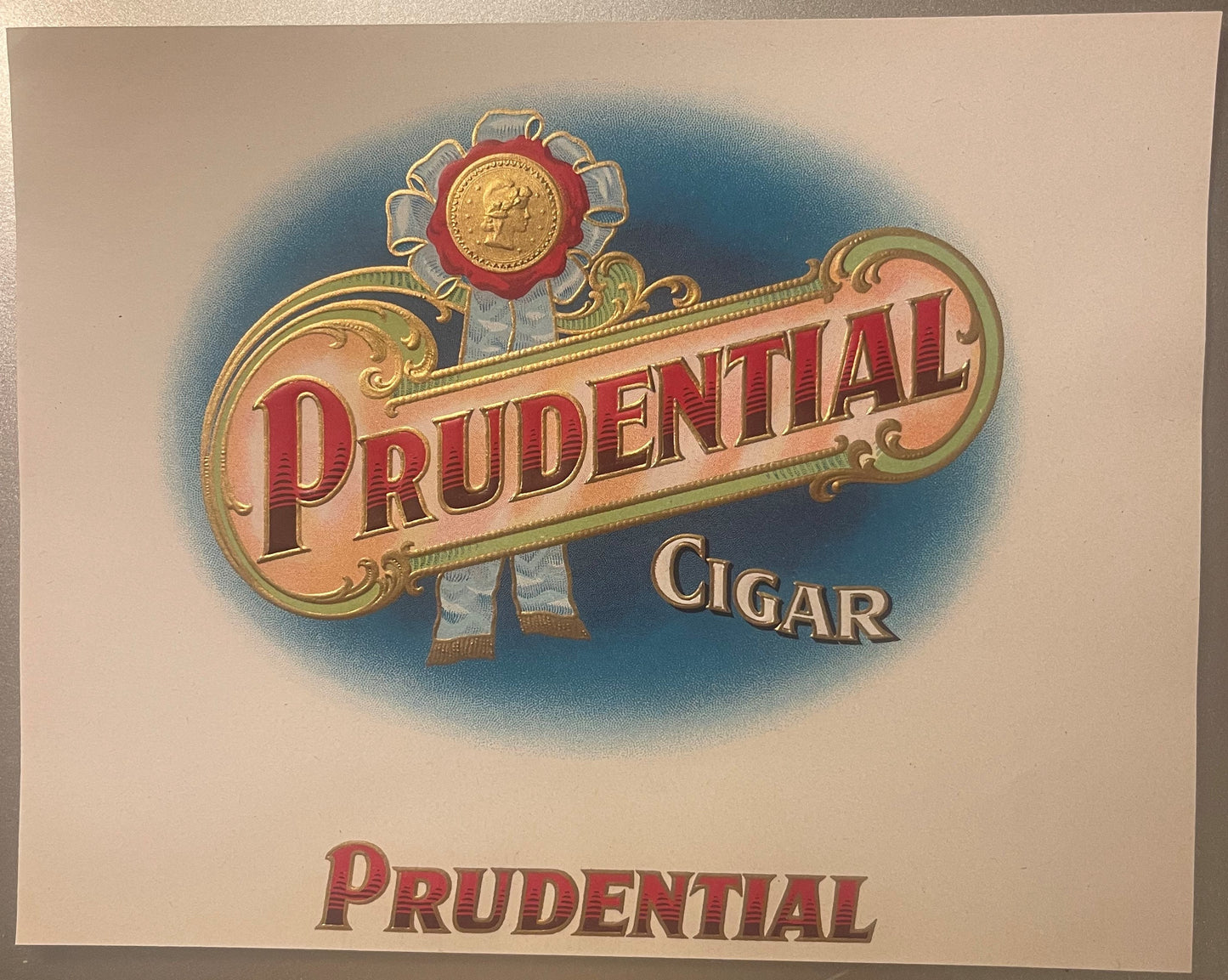 Antique embossed cigar label with "Prudential" in red letters, gold detailing, decorative medal at top, blue background, circ