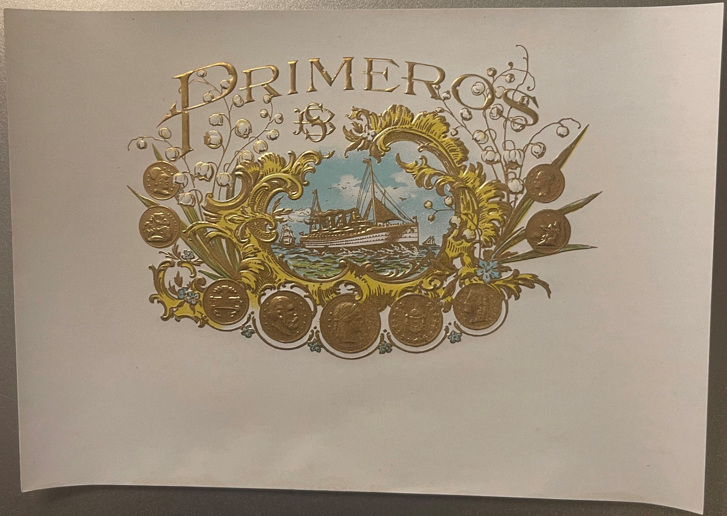 Ornate embossed gold cigar label with "Primer" text, decorative scrollwork, steamship vignette, and coin medallions on cream 